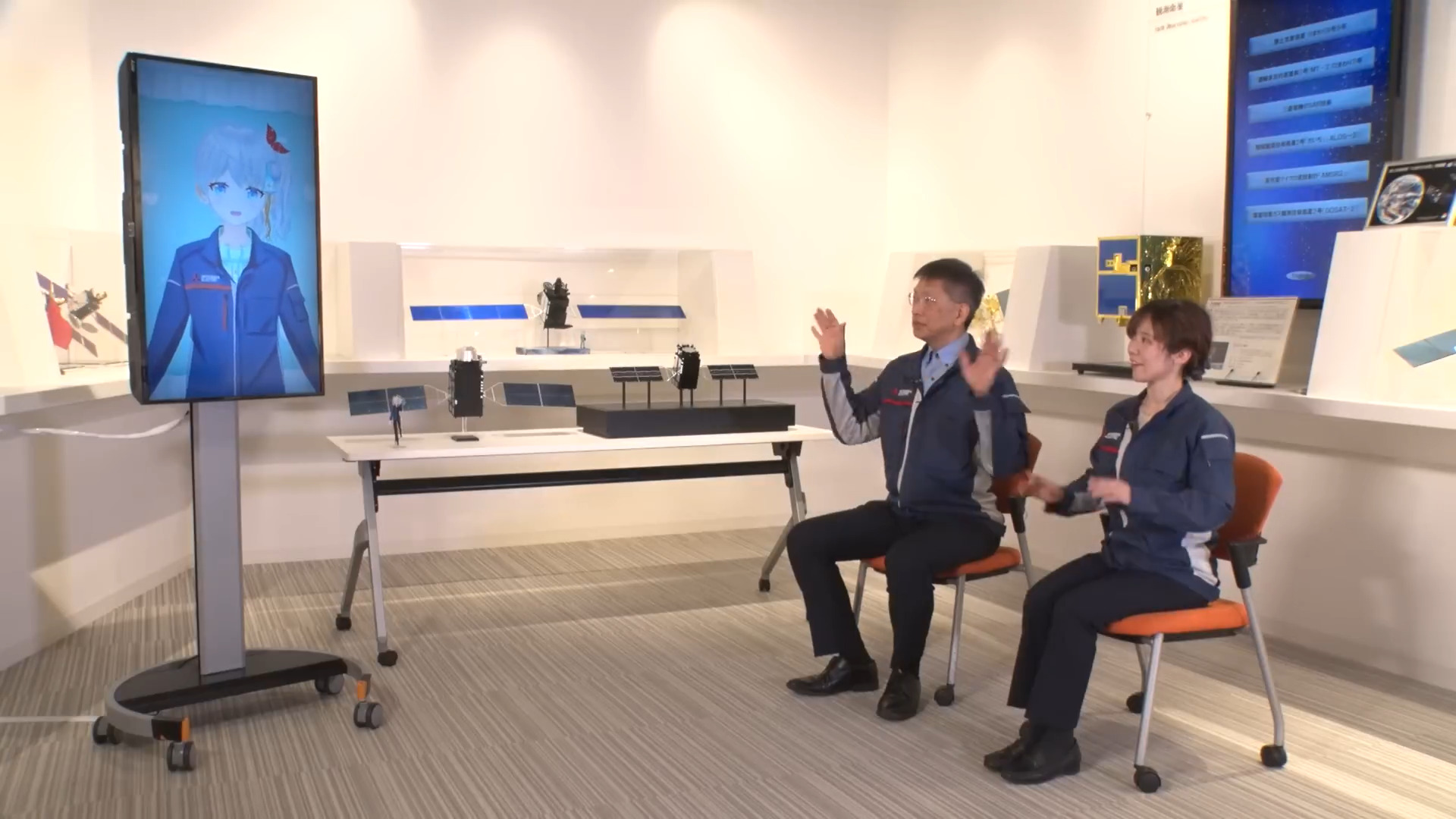 Un écran vertical affiche l'avatar animé de la VTuber Usui Kuria en uniforme bleu. Elle fait face à deux ingénieurs (un homme et une femme) vêtus de vestes de travail similaires, assis sur des chaises oranges. En arrière-plan, plusieurs maquettes de satellites équipés de panneaux solaires sont exposées sur des tables dans une pièce lumineuse.