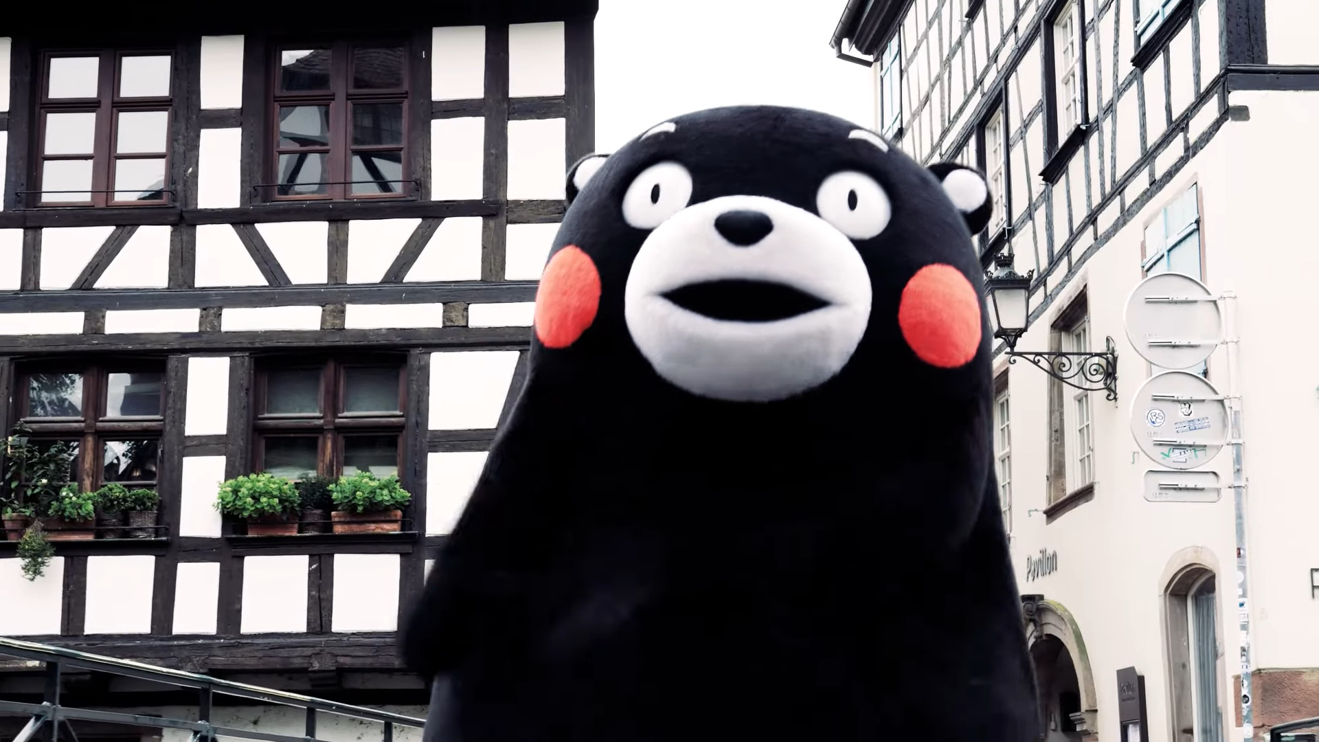 Une capture d'écran vidéo en gros plan montrant la mascotte japonaise Kumamon, un grand ours en peluche noir aux joues rouges et aux yeux écarquillés, au premier plan. Elle se tient au centre, devant une ruelle historique de Strasbourg. L'arrière-plan présente des bâtiments traditionnels à colombages (pan-de-bois) typiques, avec des plantes en pot sur les rebords de fenêtre et des réverbères anciens.